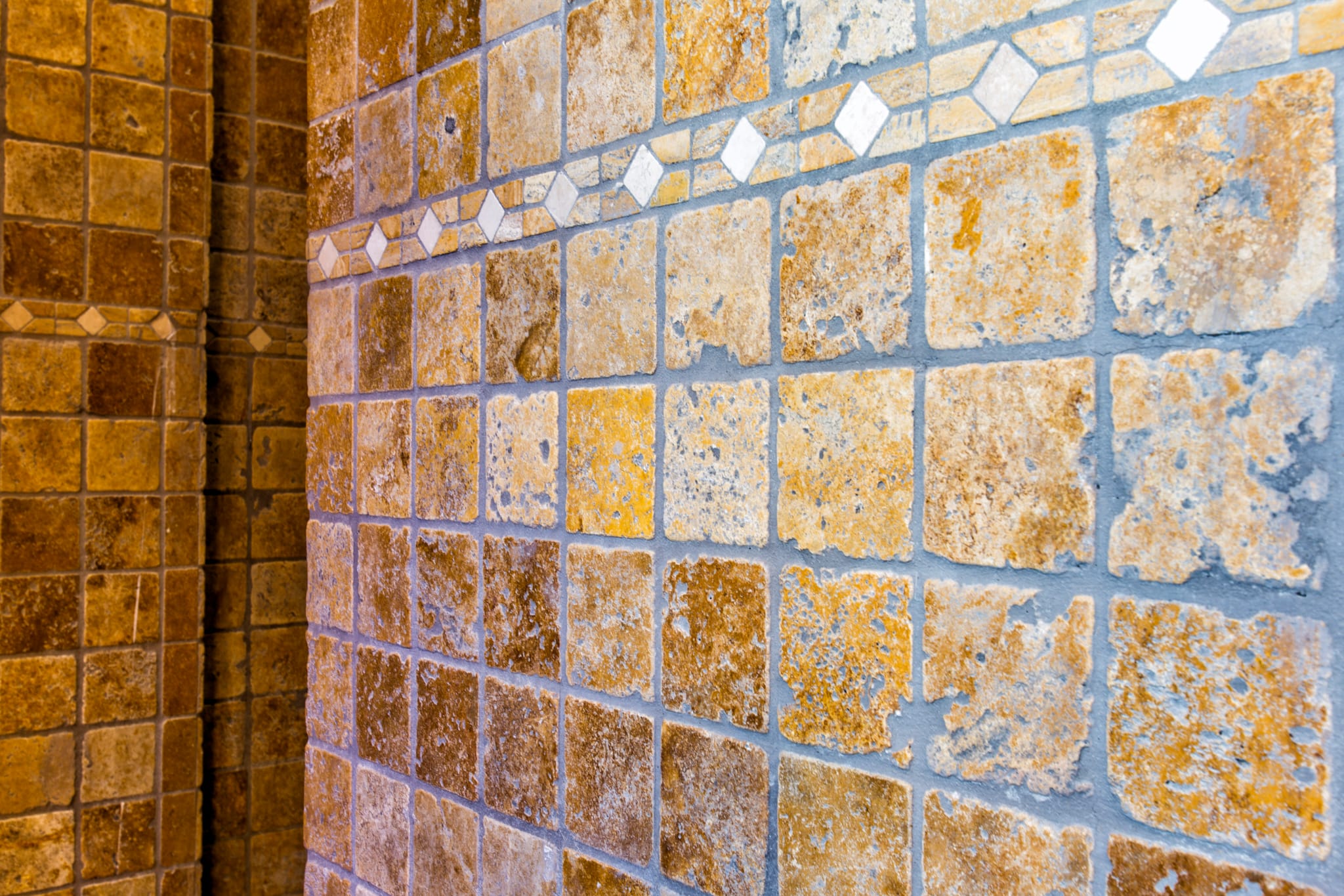 Primer plano de azulejos de piedra natural en tonos ocre con acabado rústico y una cenefa decorativa de rombos blancos.