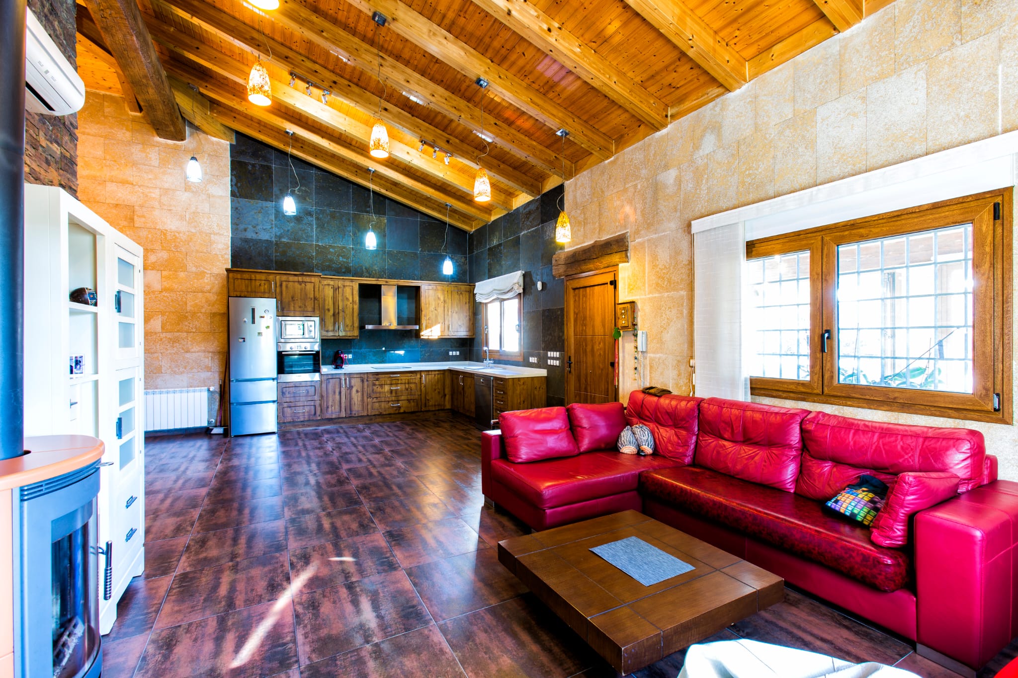 Salón-cocina diáfano con techos altos de vigas de madera, paredes de piedra, sofá de cuero rojo y cocina rústica equipada con electrodomésticos de acero inoxidable.