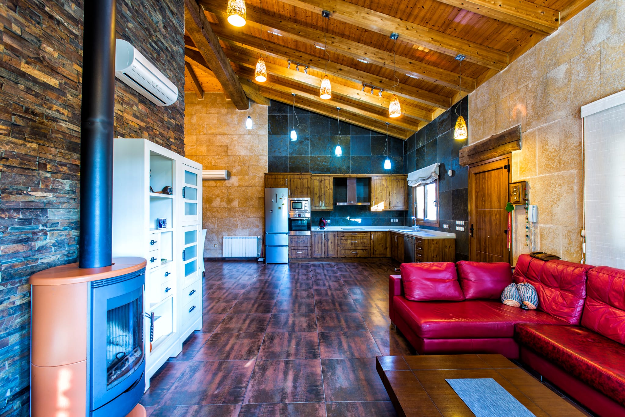 Salón-comedor diáfano con techos de vigas de madera, estufa de leña contemporánea y cocina rústica integrada bajo una pared de pizarra oscura y piedra natural.