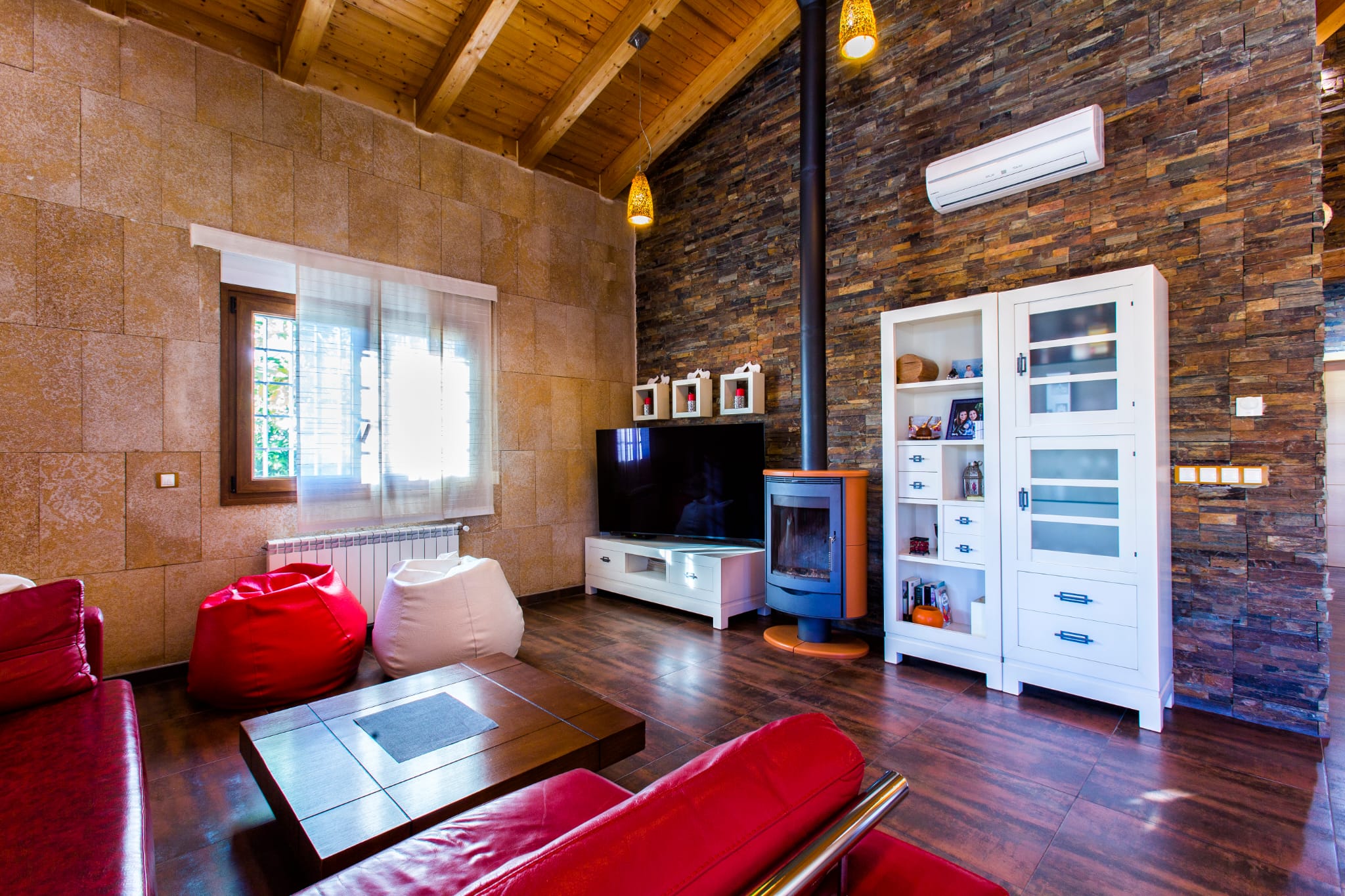Salón rústico con paredes de piedra natural, techos de vigas de madera, estufa de leña moderna y sofás de cuero rojo integrados en un ambiente cálido.