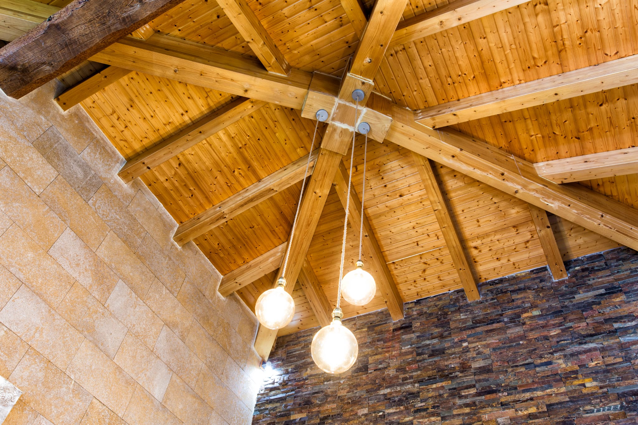 Techo abovedado con vigas de madera vistas en disposición radial, iluminado por tres lámparas colgantes esféricas sobre paredes de piedra natural y pizarra.