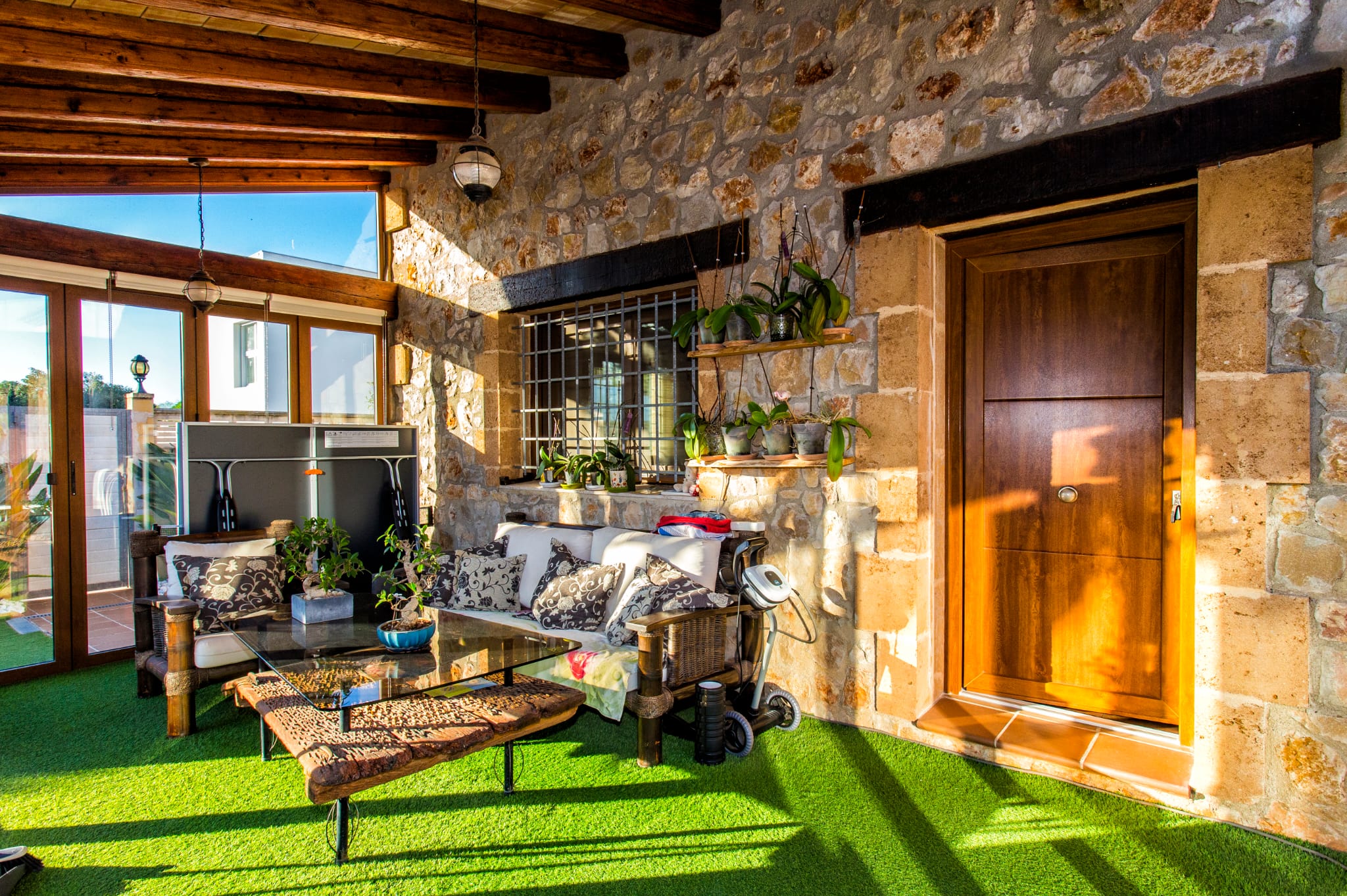 Terraza cubierta con paredes de piedra natural, vigas de madera y césped artificial. Incluye zona de estar con mesa de madera rústica y abundante luz natural.