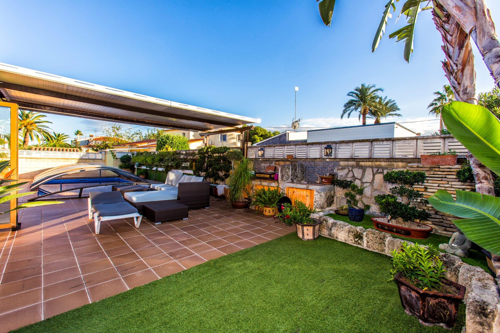 Terraza exterior con pavimentos cerámicos, césped artificial y zona de descanso bajo pérgola, junto a una piscina con cubierta telescópica y muros de piedra.