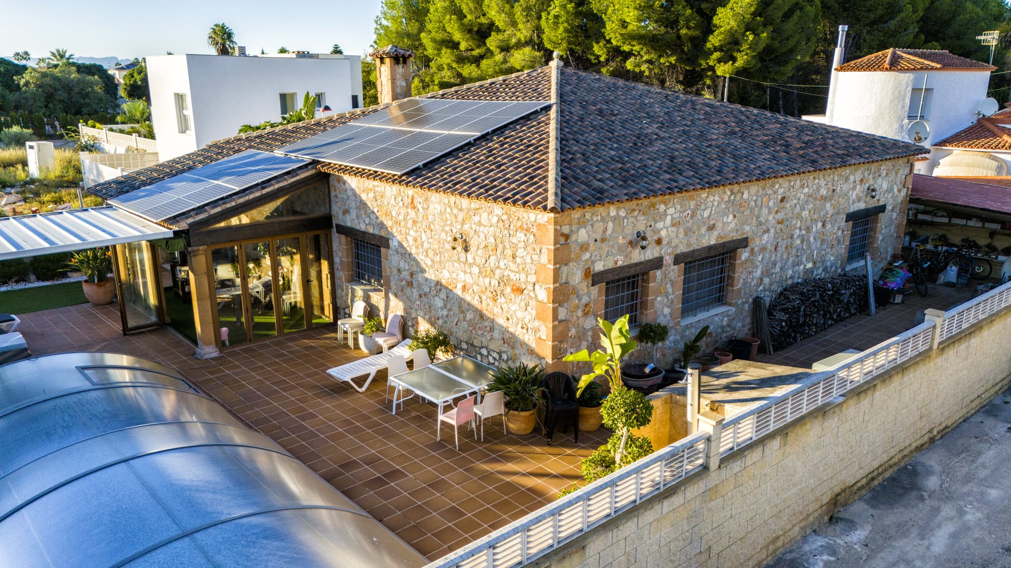 Vista exterior de una villa de piedra con paneles solares, amplia terraza de gres y piscina con cubierta curva retráctil.