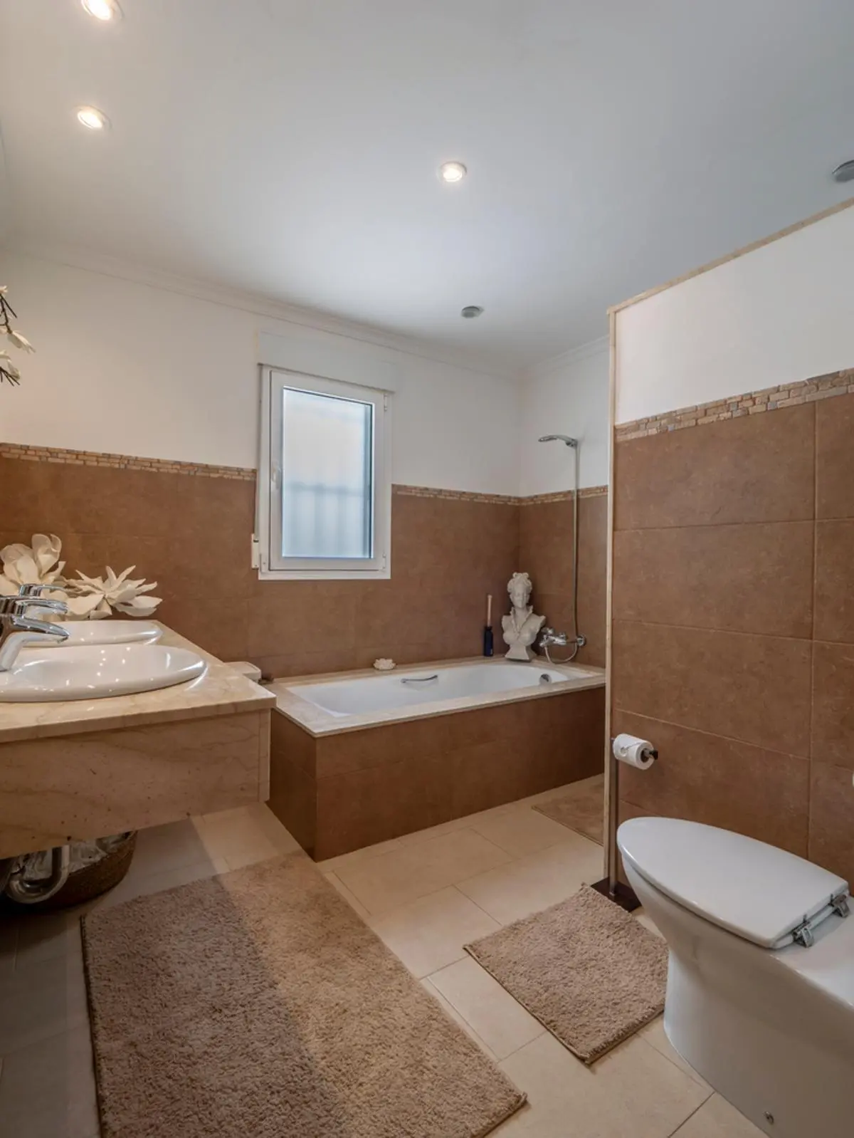 Baño completo con encimera de mármol, lavabo doble, bañera integrada revestida en gres marrón y focos empotrados en el techo.