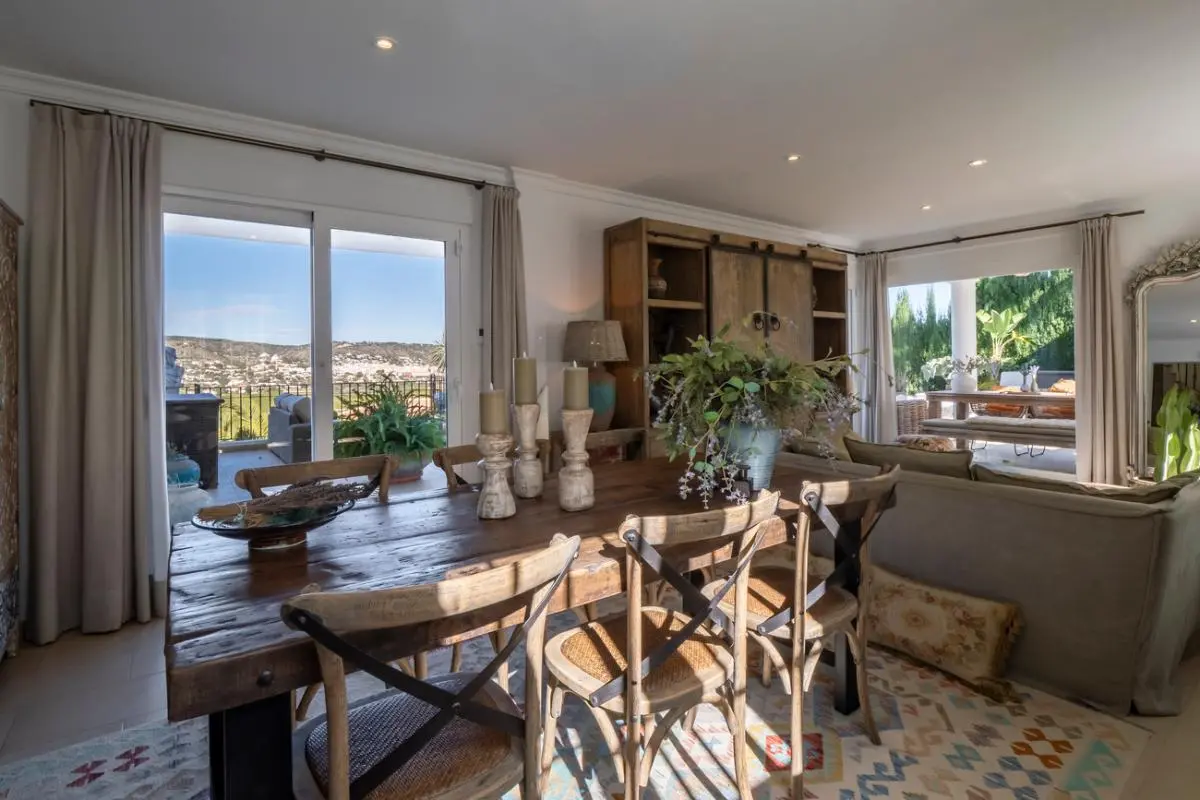 Comedor rústico con mesa de madera maciza y sillas de respaldo cruzado, con acceso directo a una terraza con vistas despejadas al valle.