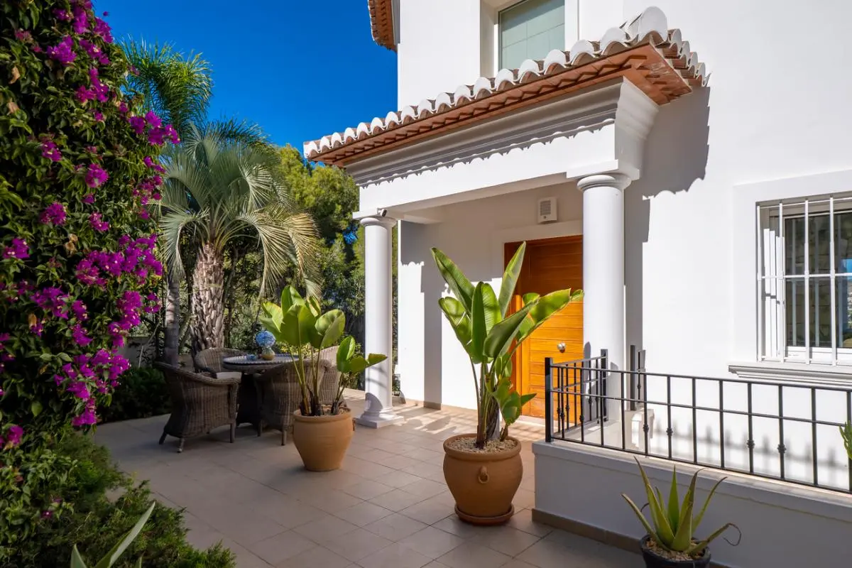 Entrada principal con porche de columnas blancas, puerta de madera y terraza pavimentada rodeada de vegetación mediterránea y buganvillas.