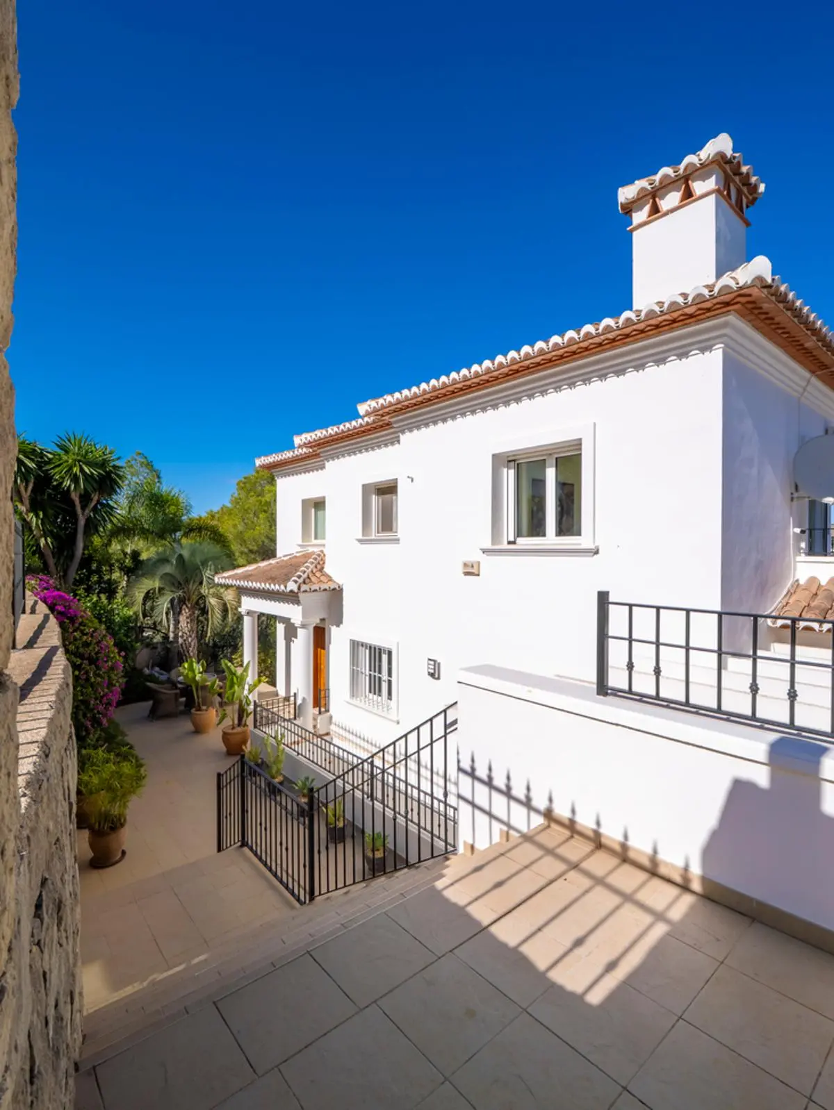 Fachada blanca de estilo mediterráneo con tejado de terracota, chimenea ornamental y barandillas de forja negra sobre una terraza pavimentada rodeada de vegetación.
