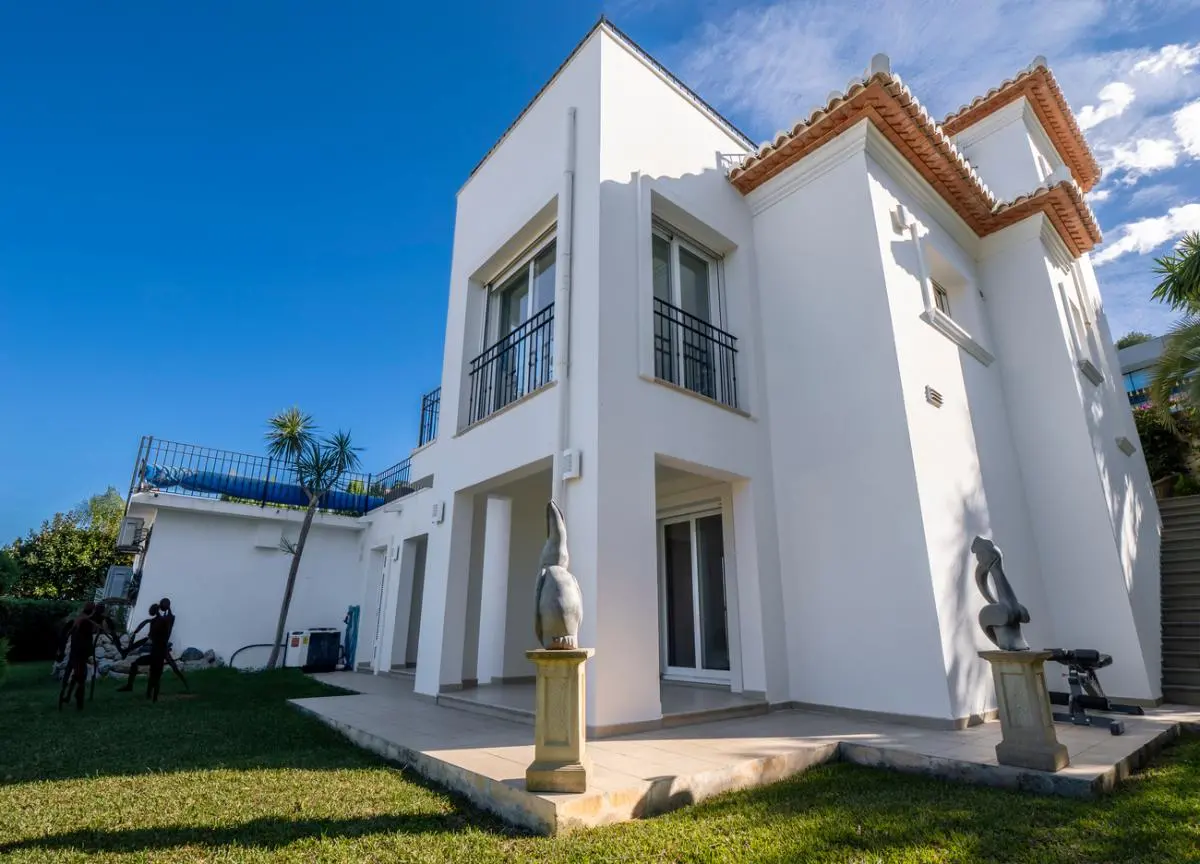 Fachada exterior de una villa blanca con tejado de teja cerámica, barandillas de hierro negro y jardín con césped y esculturas decorativas sobre pedestales.