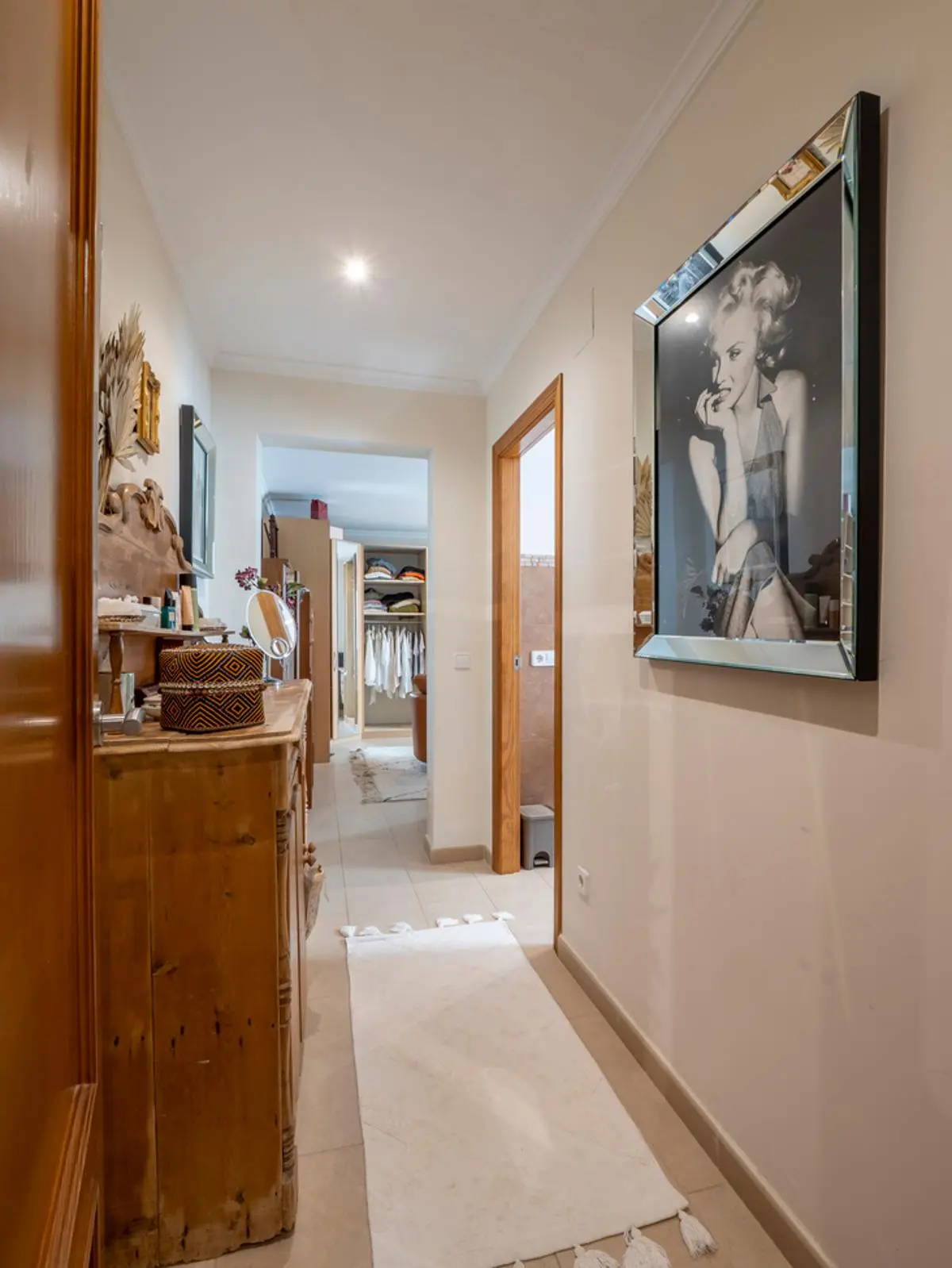Pasillo luminoso con una cómoda de madera rústica, un retrato enmarcado en espejo y acceso directo a un vestidor con estanterías y zona de colgado.