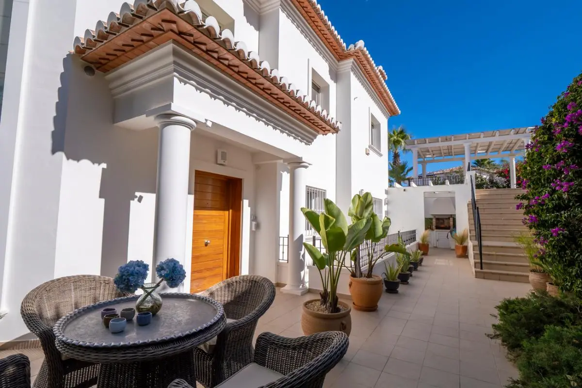 Patio exterior con zona de estar de mimbre, columnas blancas y puerta de madera. Plantas en macetas bordean el camino hacia una pérgola elevada.