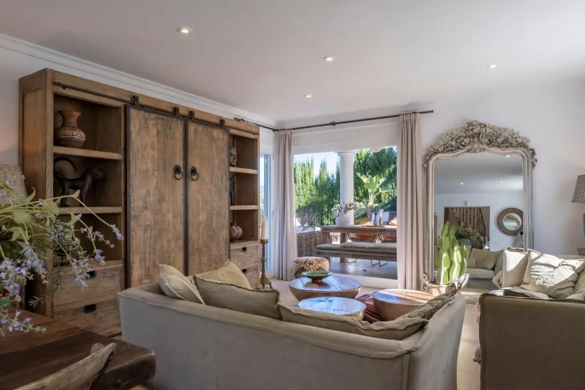 Salón con mueble rústico de madera, sofás en tonos neutros y un gran espejo ornamental, con acceso directo a una terraza con comedor exterior.