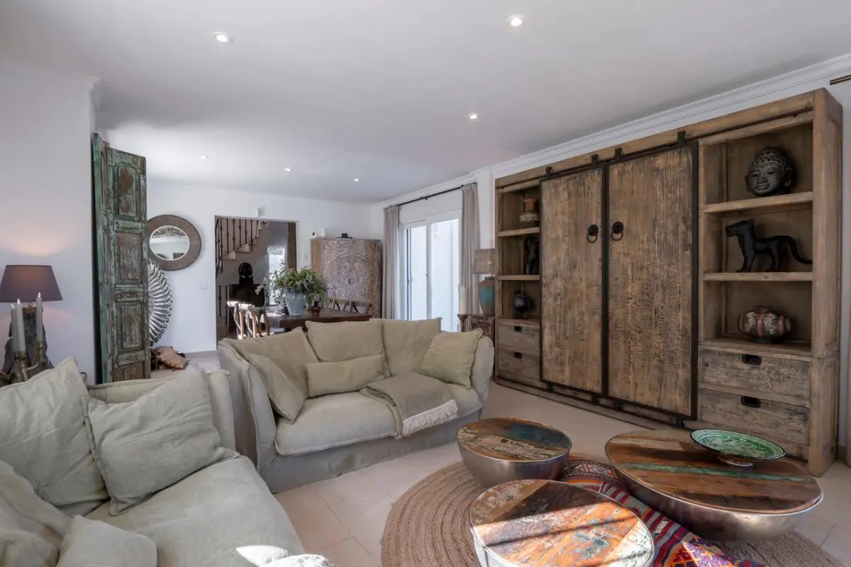 Salón con un gran mueble de madera rústica, sofás tapizados en lino beige y mesas de centro circulares de madera reciclada sobre una alfombra de yute.