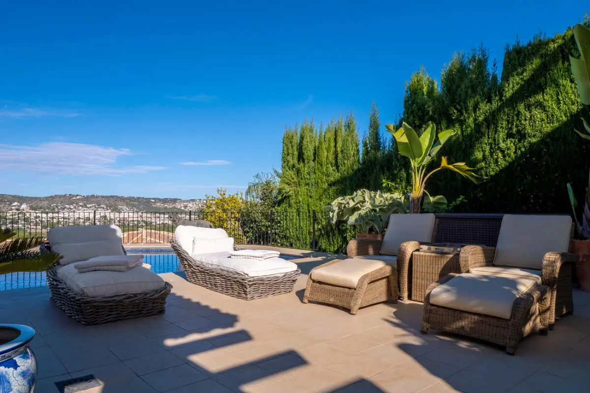 Terraza con tumbonas de mimbre y cojines blancos frente a la piscina, con vistas al valle y rodeada de cipreses y plantas tropicales.