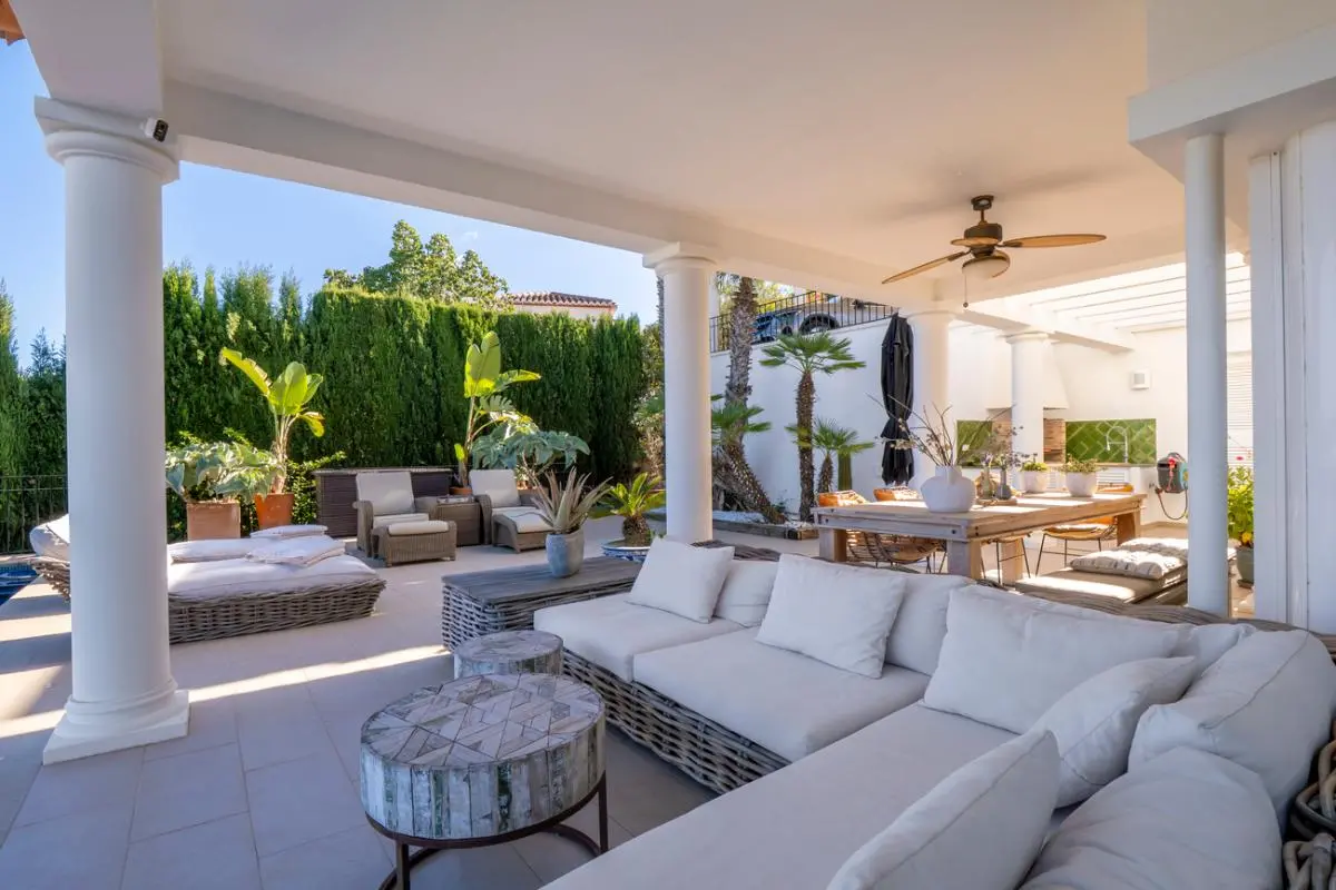 Terraza cubierta con columnas blancas, amplio sofá de mimbre gris y zona de comedor exterior con ventilador de techo, rodeada de vegetación tropical.