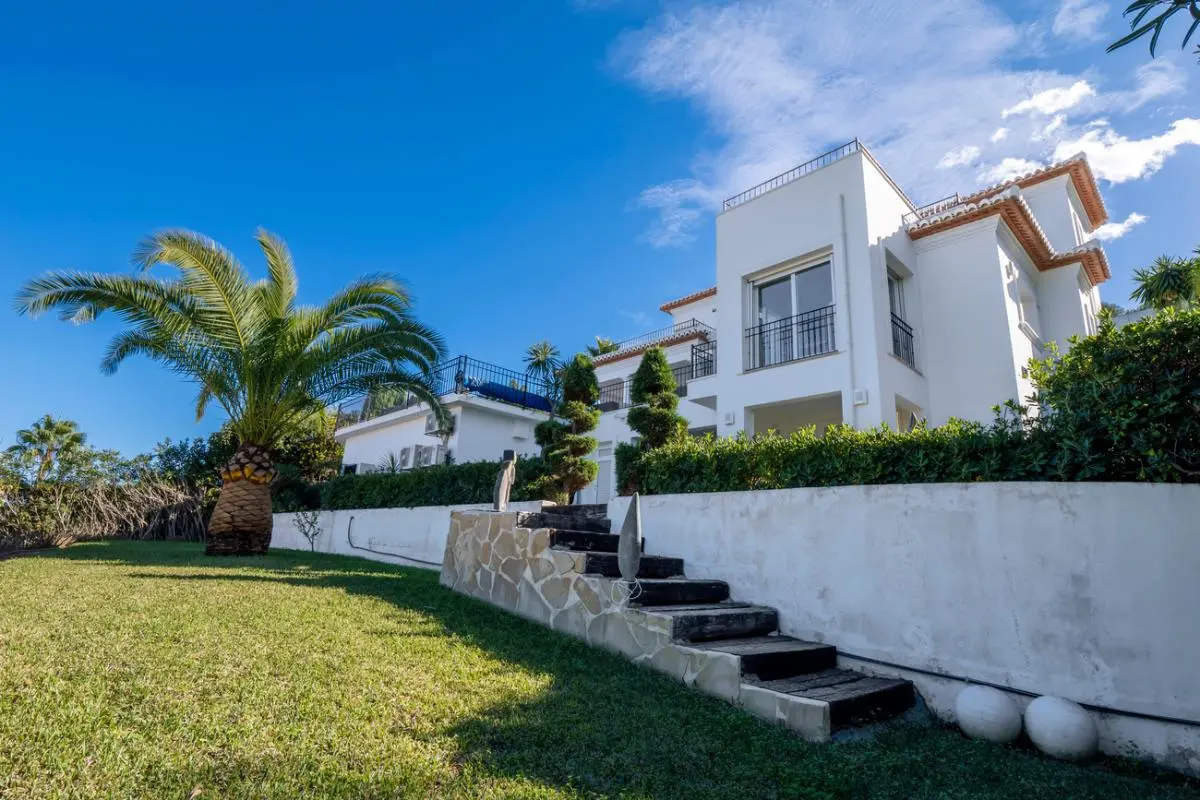 Vista exterior de una villa blanca de varios niveles con tejados de terracota, barandillas negras y un jardín con palmeras y escaleras de piedra.