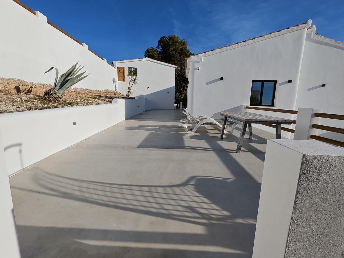 Amplia terraza exterior con suelo de microcemento, tumbonas blancas y mesa de madera, rodeada de arquitectura mediterránea de paredes blancas bajo un cielo despejado.