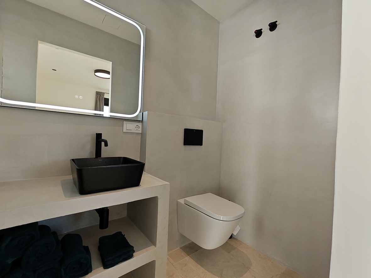 Baño de diseño minimalista con lavabo sobre encimera negro, espejo retroiluminado y sanitario suspendido sobre paredes de microcemento.