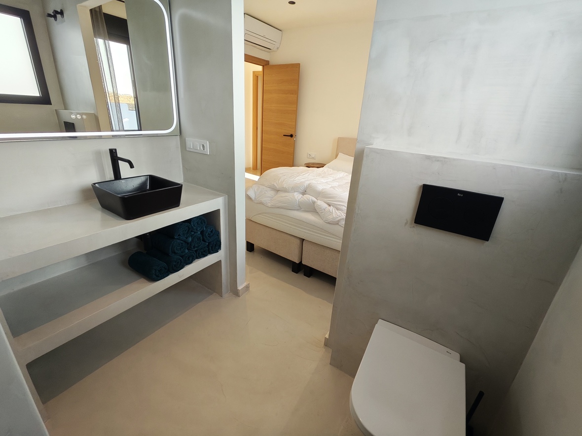 Baño en suite de estilo industrial con acabados de microcemento, lavabo de sobreponer negro y espejo retroiluminado conectado al dormitorio.
