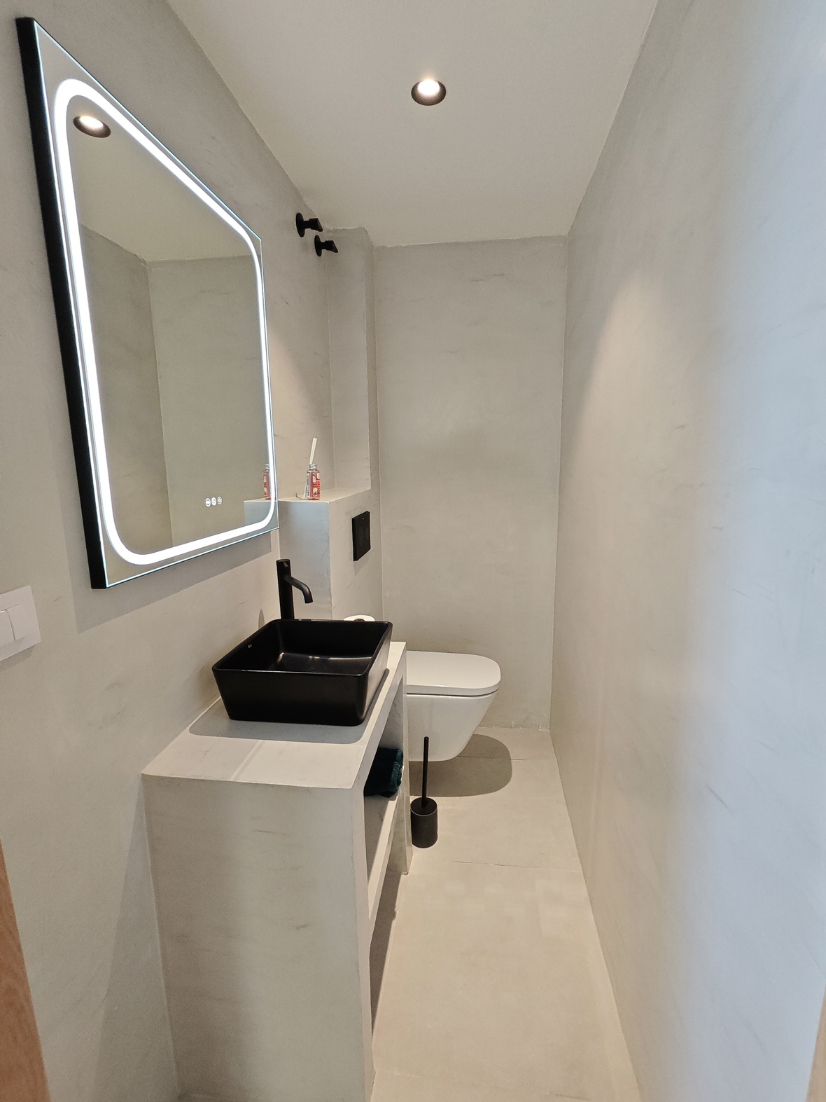 Baño minimalista con lavabo sobre encimera negro, grifería mate, espejo retroiluminado LED y paredes de microcemento gris claro.