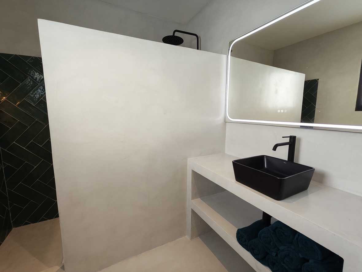 Baño moderno con acabados de microcemento, lavabo sobre encimera negro y espejo retroiluminado. La ducha abierta destaca por sus azulejos en espiga verde oscuro y grifería negra.