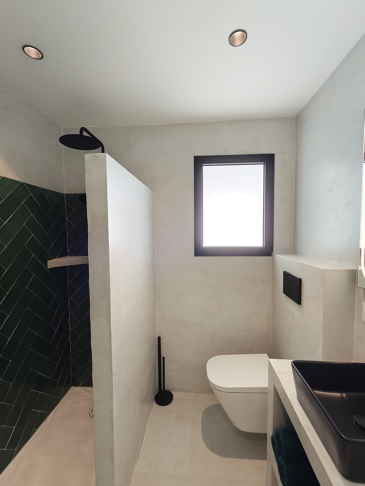 Baño moderno con ducha de azulejos verdes en espiga, inodoro suspendido y paredes de microcemento blanco con grifería negra.