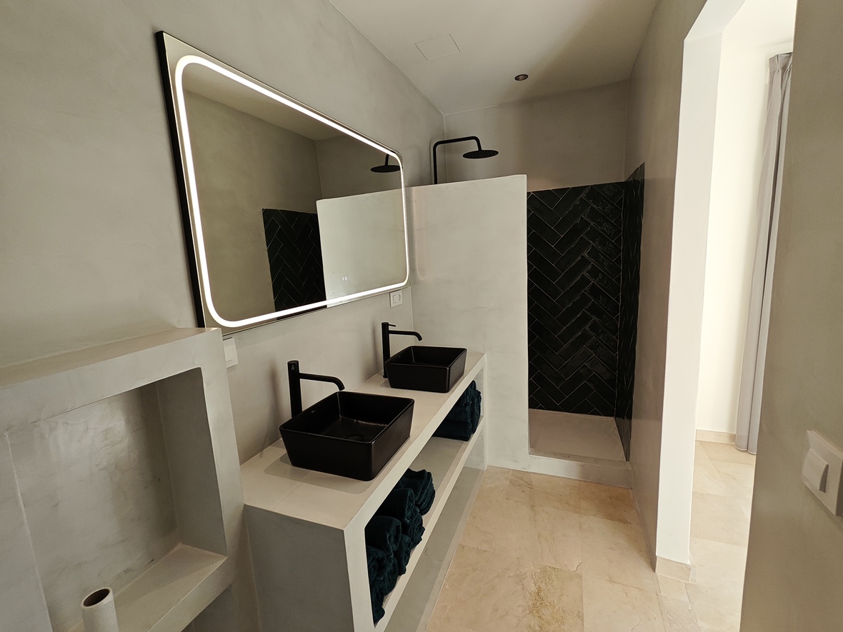 Baño moderno con lavabo doble de microcemento, lavabos negros sobre encimera, espejo retroiluminado y ducha con azulejos negros en espiga.