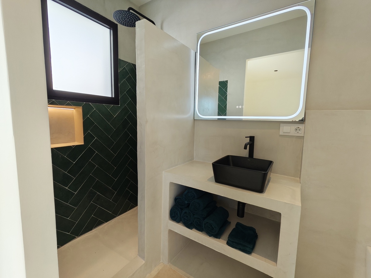 Baño moderno con lavabo sobre encimera negro, espejo con retroiluminación LED y ducha con azulejos verdes en espiga y paredes de microcemento.