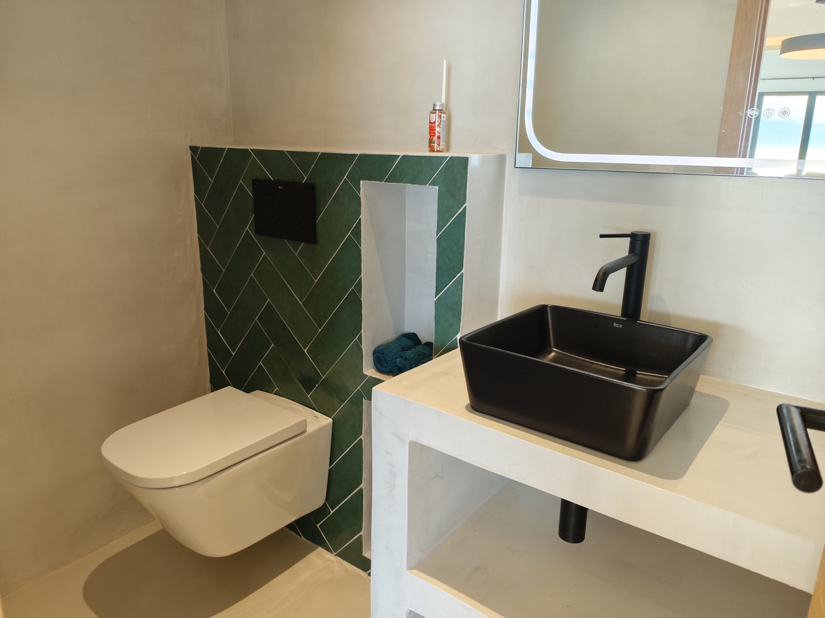Baño moderno con lavabo sobre encimera negro, paredes de microcemento y un detalle de azulejos verdes en espiga junto al inodoro suspendido.