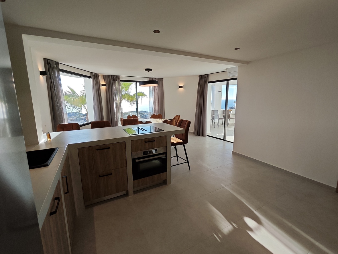 Cocina abierta con armarios de madera, encimera gris e isla con placa de inducción, conectada a un comedor con vistas al mar y acceso directo a la terraza.