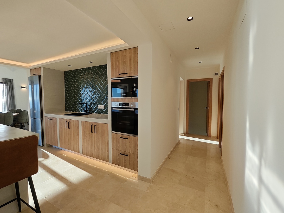 Cocina moderna con armarios de madera, salpicadero de azulejos verdes en espiga y electrodomésticos integrados, junto a un pasillo con suelo de piedra pulida.