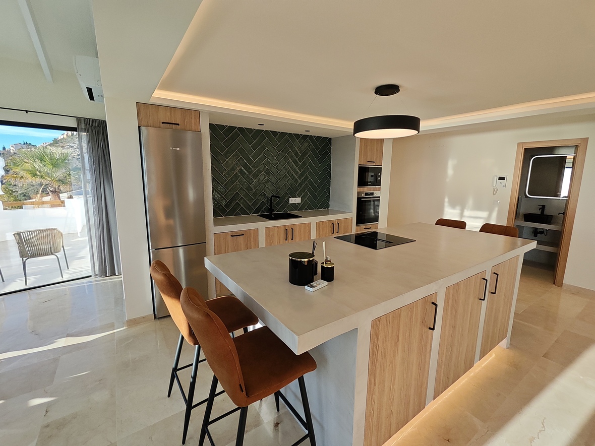 Cocina moderna con isla central, encimera de piedra, taburetes de cuero y salpicadero de azulejos verdes en espiga. Incluye electrodomésticos integrados y acceso a terraza.
