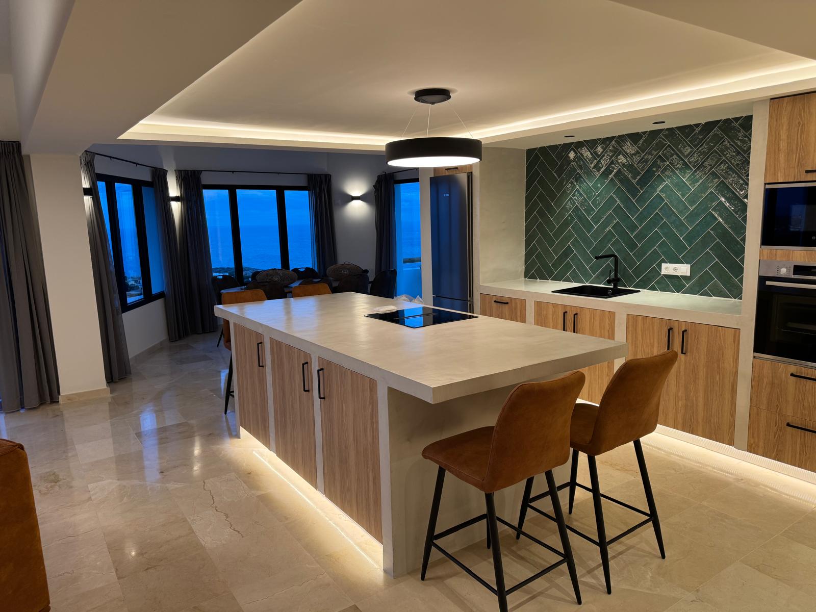 Cocina moderna con isla de microcemento, salpicadero de azulejos verdes en espiga y vistas panorámicas al mar desde los ventanales durante el anochecer.
