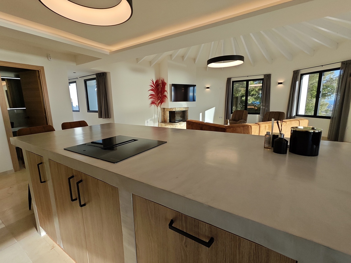 Cocina moderna de concepto abierto con isla de encimera clara y placa de inducción, integrada a un salón con techos de vigas blancas y chimenea de piedra.