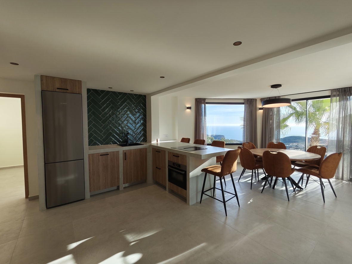 Cocina moderna de planta abierta con salpicadero de espiga verde, electrodomésticos integrados y zona de comedor con vistas al mar y luz natural.