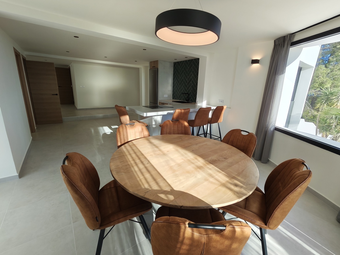 Comedor de planta abierta con mesa redonda de madera, sillas color teja y lámpara circular, integrado con una cocina moderna de salpicadero en espiga.