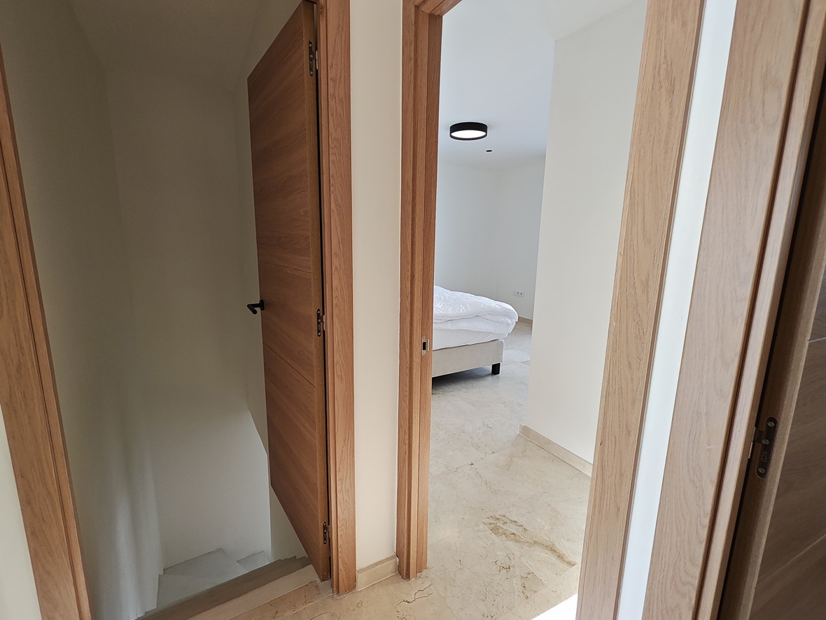 Distribuidor con puertas de madera de roble y suelos de piedra natural, que conecta el acceso a un dormitorio con la escalera hacia la planta inferior.