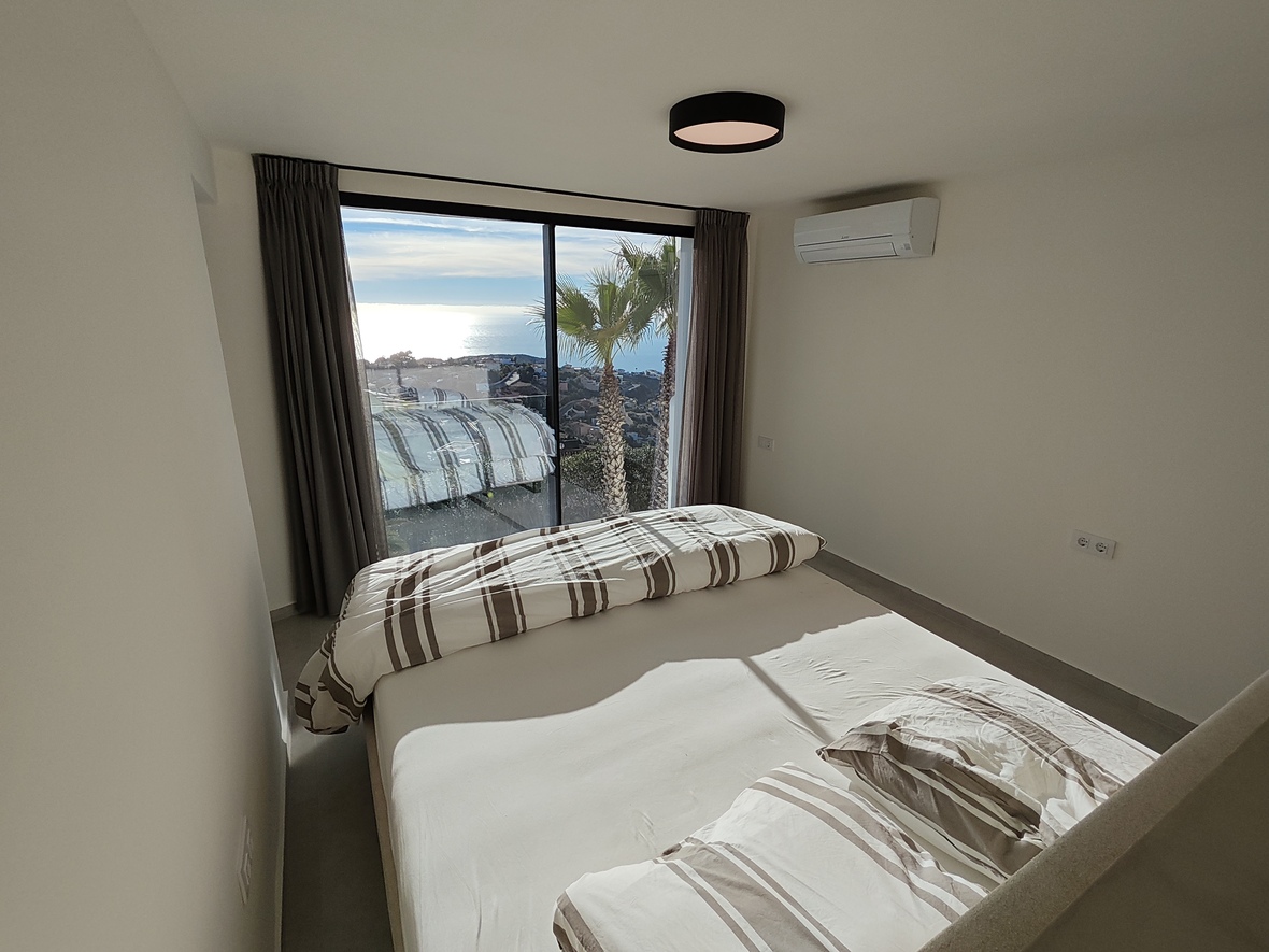 Dormitorio con ventanales de suelo a techo con vistas al mar y palmeras. Incluye paredes blancas minimalistas, ropa de cama a rayas y aire acondicionado de pared.