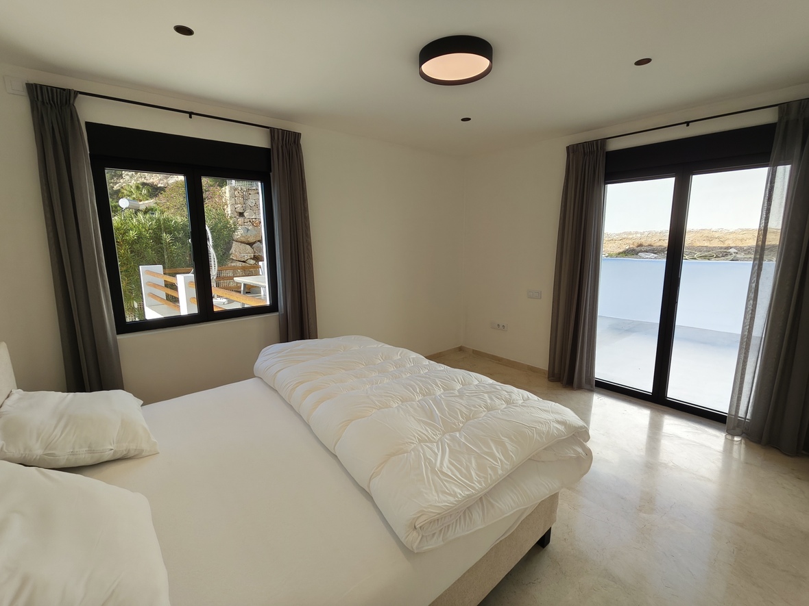 Dormitorio luminoso con suelo de mármol, ventanales de marco negro con acceso a terraza y cortinas grises. Incluye iluminación empotrada y plafón minimalista.