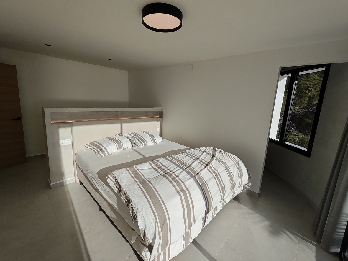 Dormitorio moderno con cabecero de obra divisorio e iluminación LED integrada, suelos de gres porcelánico y ventanal con marco negro hacia el exterior arbolado.