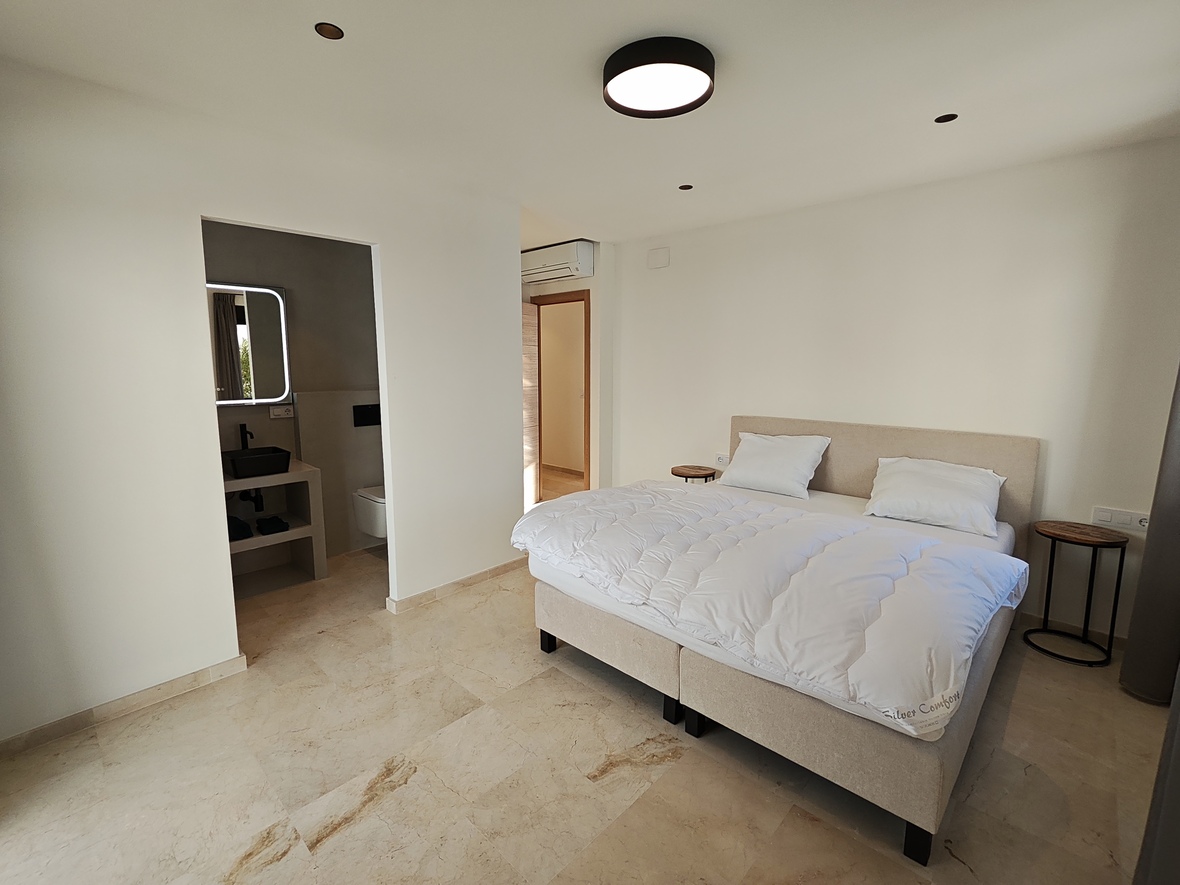 Dormitorio moderno con suelos de mármol, cama doble tapizada y baño integrado que incluye un lavabo de sobremesa negro y espejo con iluminación LED.