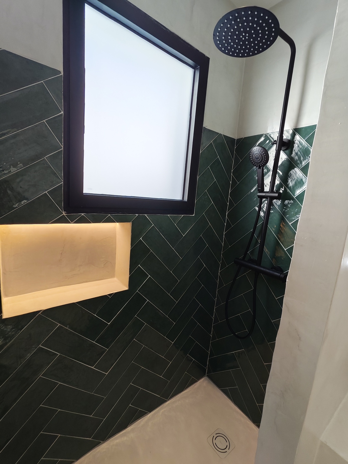 Ducha moderna con azulejos verdes en espiga, grifería negra mate, hornacina iluminada y ventana de cristal translúcido que aporta luz natural y privacidad.
