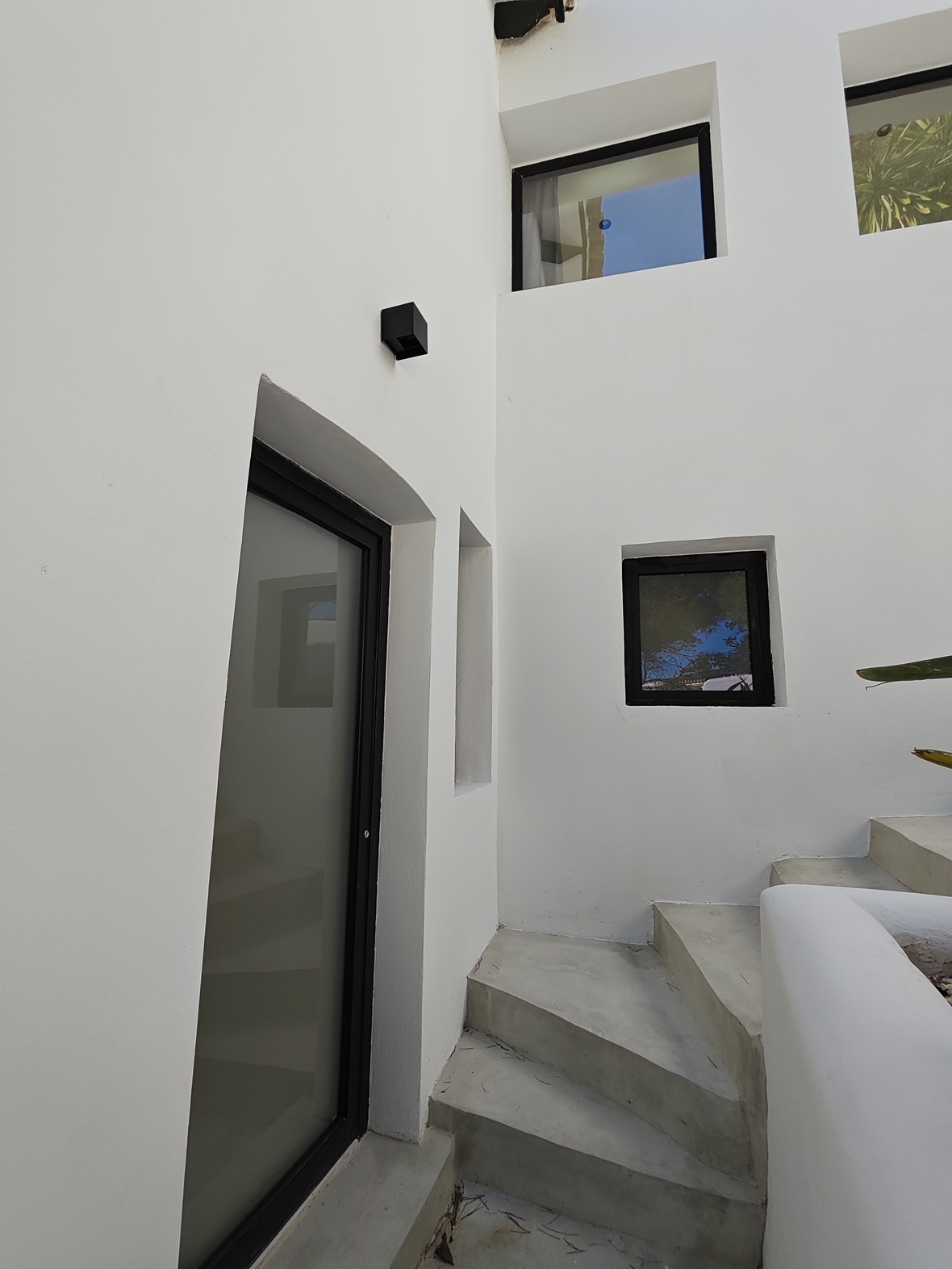 Escalera exterior de hormigón integrada en muros blancos minimalistas, con ventanas de marco negro y una puerta de cristal traslúcido.