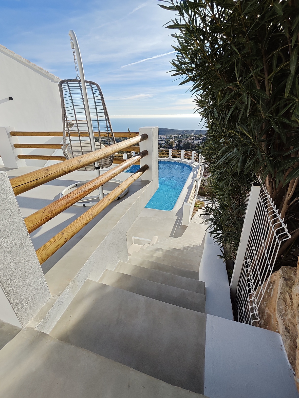 Escaleras de hormigón acceden a una terraza con silla colgante y barandillas de madera, con vistas a la piscina y al horizonte del mar Mediterráneo.