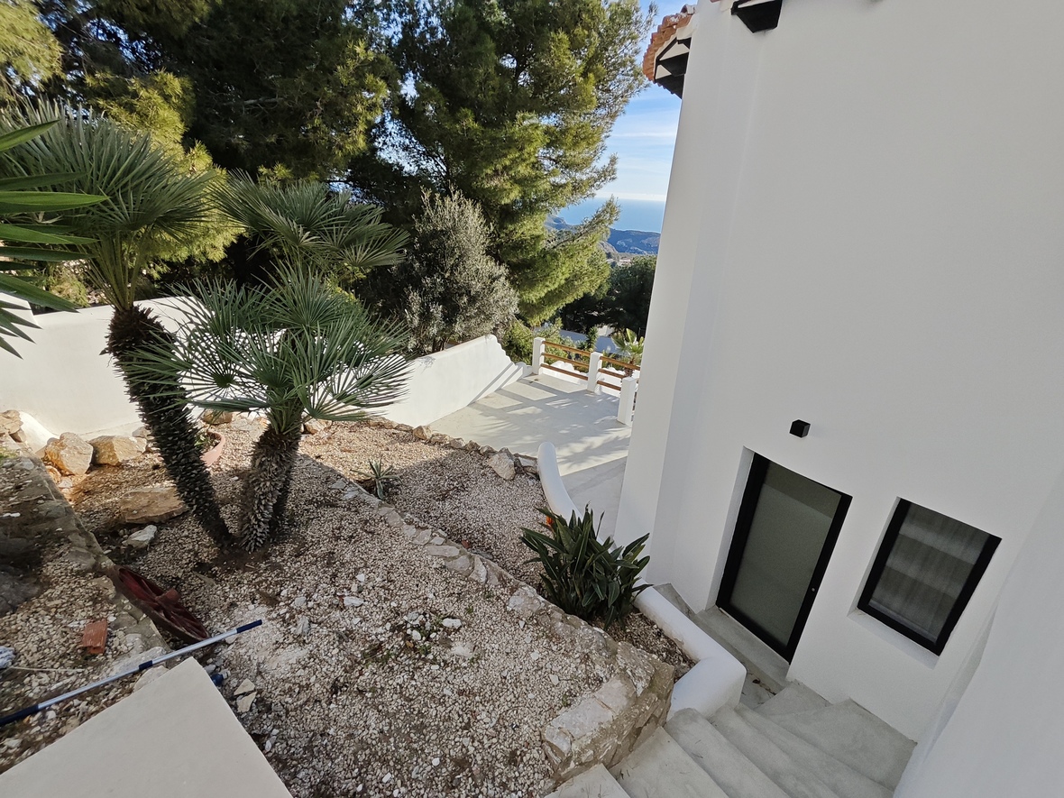 Exterior de la villa con jardín de gravilla y palmeras, muros de piedra y escaleras de hormigón que bajan hacia una entrada negra con vistas al mar.