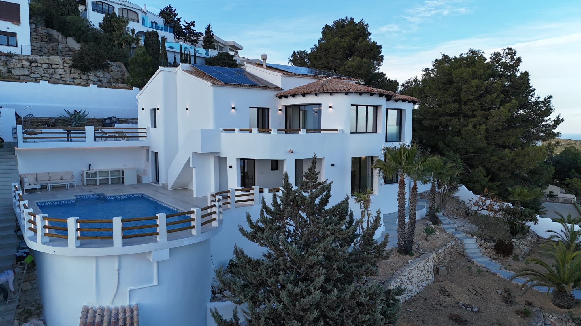 Fachada de una villa blanca con piscina curva, paneles solares integrados y jardines aterrazados de piedra natural con palmeras y pinos.