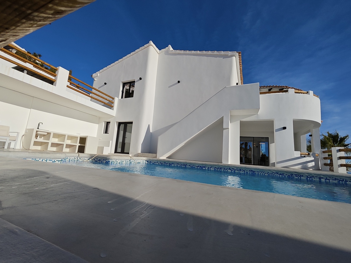 Fachada de villa blanca con piscina de gresite azul, cocina exterior integrada y barandillas de madera bajo un cielo despejado.