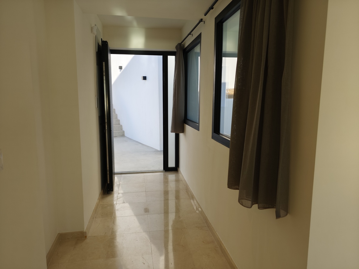 Pasillo con suelo de mármol pulido que da acceso a un patio exterior con escaleras. Presenta carpintería negra y cortinas de lino en tonos tierra.