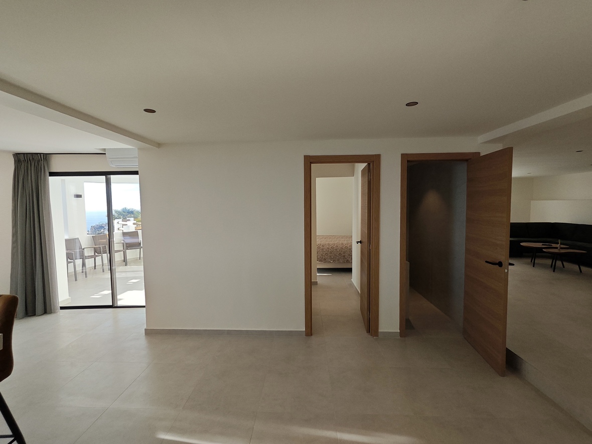 Pasillo interior con suelos cerámicos claros, carpintería de madera natural y acceso acristalado a una terraza con vistas panorámicas al mar.