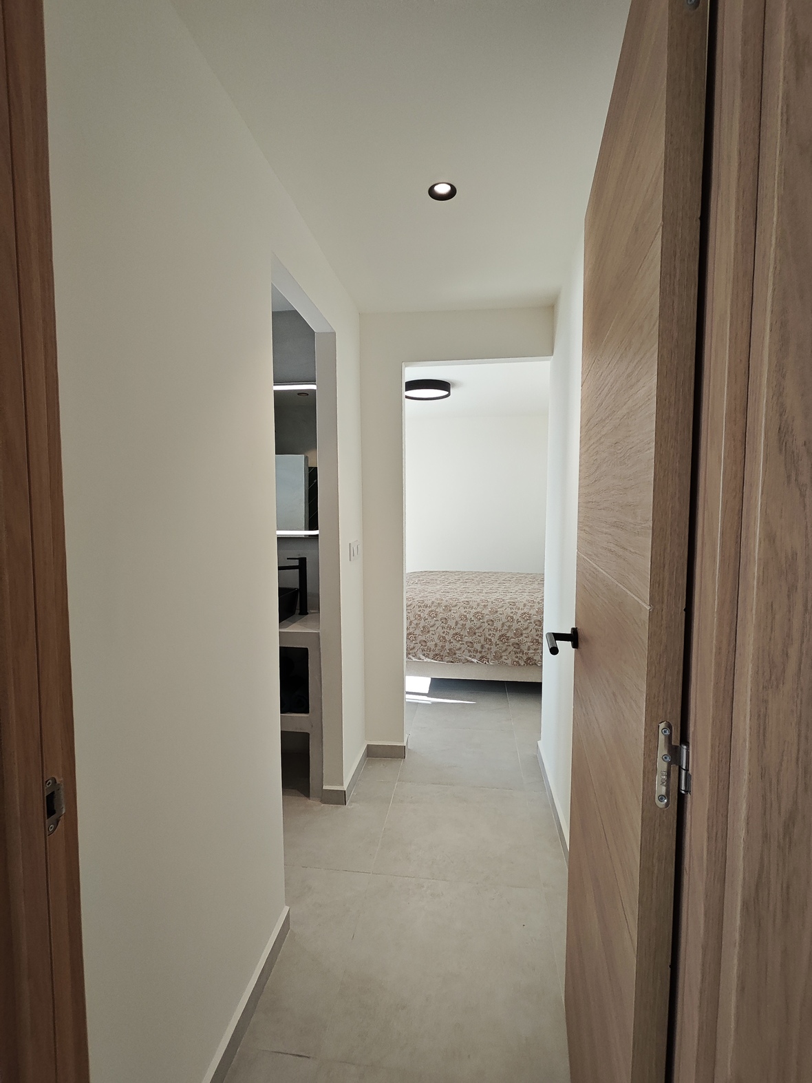 Pasillo moderno con puertas de madera clara y suelo porcelánico gris, que conecta un baño de diseño minimalista con un dormitorio luminoso.